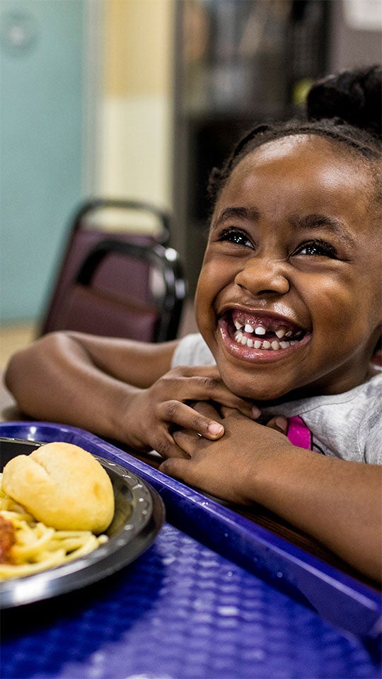 A child sits at a table with a blue tray holding a plate of spaghetti, sauce, and a bread roll, hands clasped together.